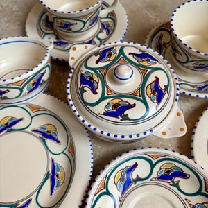 Puede incluir: Un juego de vajilla de cerámica color crema con un diseño geométrico azul, verde y amarillo. El juego incluye platos, cuencos, tazas, platillos y una fuente para servir con tapa. Los bordes están decorados con puntos azules.