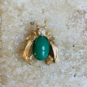 May include: A gold-toned brooch in the shape of a beetle with a green oval cabochon stone and a white pearl accent.