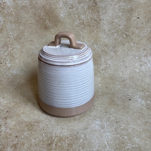 May include: A ceramic jar with a lid. The jar is white with horizontal ridged detailing and a tan base. The lid is white with a tan handle. The jar is designed for kitchen or home decor.