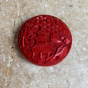 May include: A round, red carved lacquer box featuring a detailed Asian landscape scene. The design includes a multi-tiered pagoda, mountains, trees, and a crescent moon. The intricate carving is highlighted by the vibrant red color.