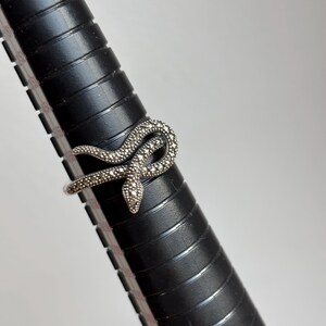 May include: A silver-toned ring in the shape of a snake, adorned with small, sparkling stones. The snake is coiled, with its head facing forward. The ring is displayed on a black ring sizer.