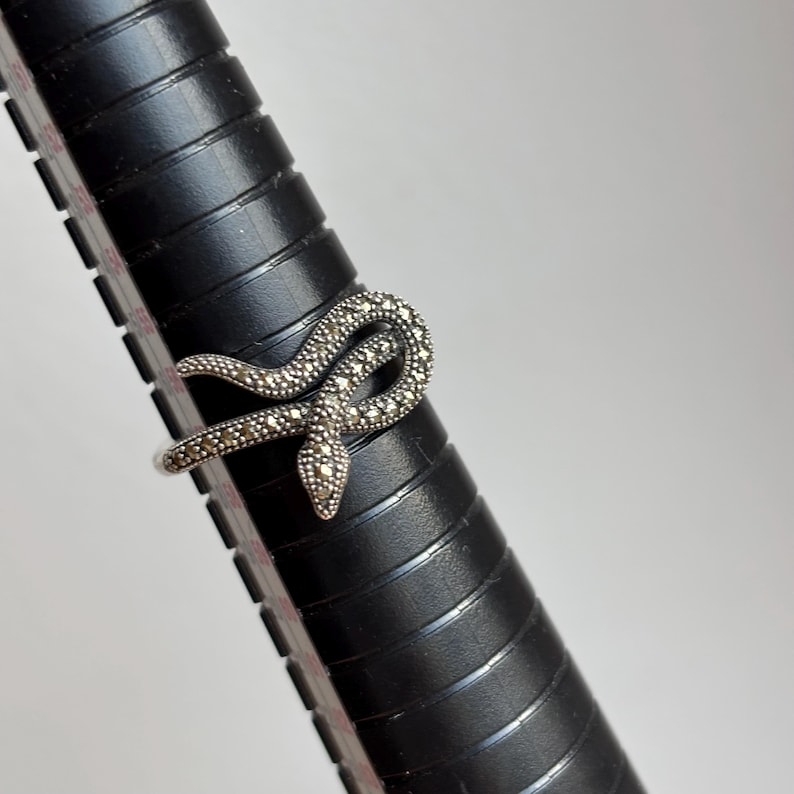 May include: A silver-toned ring in the shape of a snake, adorned with small, sparkling stones. The snake is coiled, with its head facing forward. The ring is displayed on a black ring sizer.