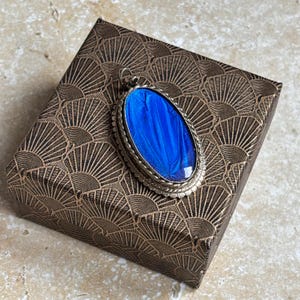 May include: A silver pendant with a bright blue, oval-shaped inlay, set on a decorative brown box with a repeating fan pattern. The pendant's vivid blue colour contrasts with the box's warm tones, creating a visually appealing composition.