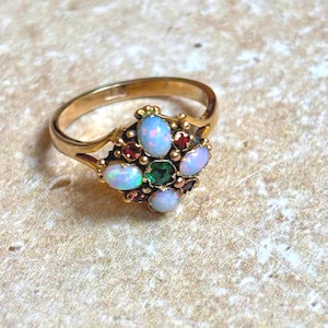 Op de afbeelding: Een gouden ring met een cluster van edelstenen. De ring heeft ovale opalen, een centrale smaragd en kleine rode granaatstenen. De ring heeft een sierlijk ontwerp met gedetailleerde kenmerken.