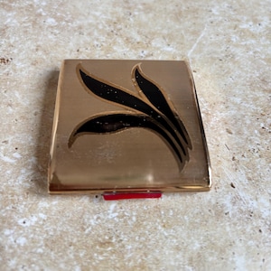 Op de afbeelding: Een vintage goudkleurige compactpoederdoos met een zwart bladerontwerp. De vierkante compactpoederdoos heeft een geribbelde textuur en een rode sluiting. Het zwarte bladerontwerp is gecentreerd op het deksel van de compactpoederdoos.