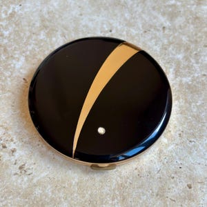 Op de afbeelding: Een ronde, zwarte compactpoederdoos met een goudkleurige diagonale streep en een klein, rond, zilverkleurig accent. De compactpoederdoos heeft een goudkleurige rand en een kleine sluiting.