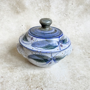 Könnte beinhalten: Eine weiße, überdachte Keramikschale mit einem blauen und grünen Blumendesign. Der Deckel hat einen kleinen Knopf oben.