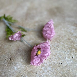 Może przedstawiać: Trzy różowe jedwabne kwiaty maku z żółtymi środkami. Kwiaty są ułożone na jasnobrązowej, teksturowanej powierzchni.