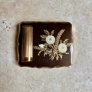 May include: A vintage brown compact with a gold-tone lipstick and floral design. The compact has a scalloped edge and features a gold floral design with white and light blue accents. The lipstick is gold and cylindrical.