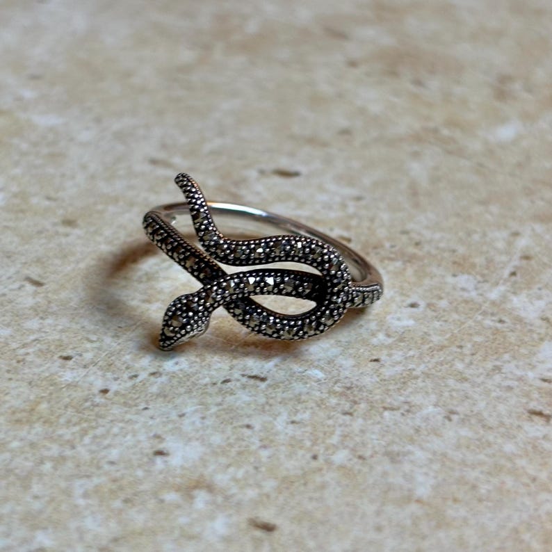 May include: A silver-toned ring featuring a snake design. The snake is coiled and detailed with small, dark stones, creating a textured appearance. The ring is set against a neutral background.