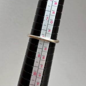 May include: A silver ring is placed on a black ring sizer. The ring is round with a smooth, shiny surface. The ring sizer has white markings and numbers for measuring ring sizes.