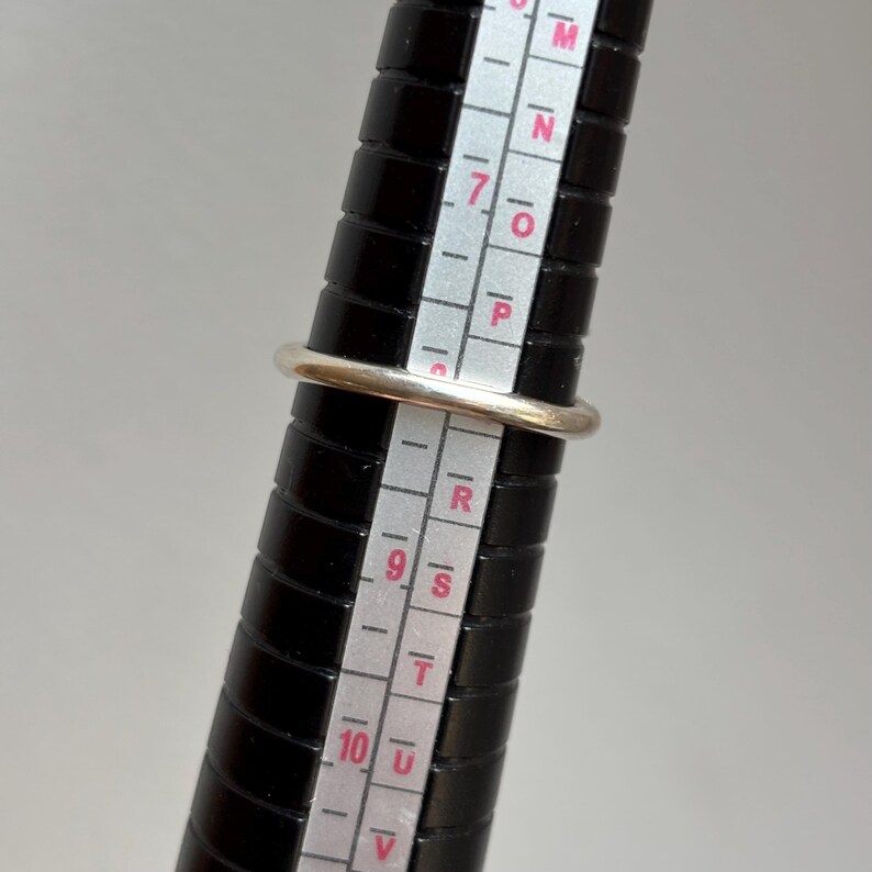 May include: A silver ring is placed on a black ring sizer. The ring is round with a smooth, shiny surface. The ring sizer has white markings and numbers for measuring ring sizes.