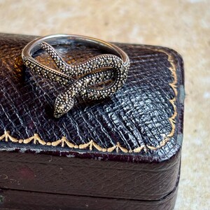 May include: A silver-toned ring in the shape of a snake, encrusted with small, sparkling stones. The snake coils around the finger, with its head raised. The ring is displayed on a dark, textured surface.