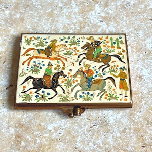 May include: A rectangular metal compact case with a hinged lid featuring a colorful illustration of four figures on horseback playing polo. The scene includes floral motifs, green trees, and a standing figure on a cream background with gold trim.