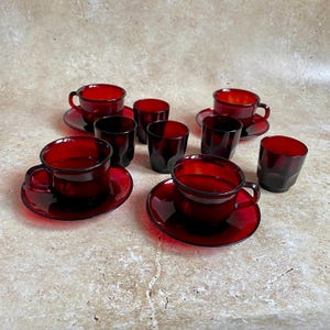 Op de afbeelding: Een verzameling vintage robijnrode glazen theekopjes met bijpassende schotels en kleine bekers. De kopjes hebben oren en staan op ronde schotels. De bekers zijn kort en cilindrisch. Het glaswerk is gerangschikt op een neutraal oppervlak.