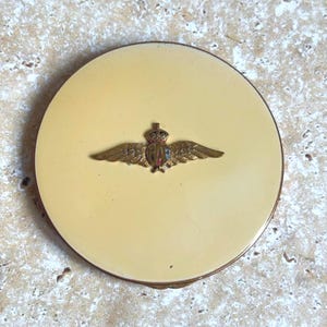 May include: A round, cream-coloured compact case with a metallic gold-tone rim. Centered on the lid is a brass-coloured emblem featuring a crown resting above a shield flanked by two outstretched wings, with small red and blue enamel accents.