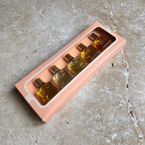 May include: A fragrance collection set in a pink box. The box contains five small perfume bottles with gold caps. The bottles are filled with various shades of amber-colored liquid. The box is labeled "FRAGRANCE COLLECTION".