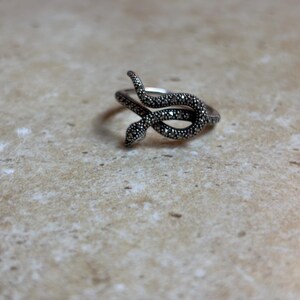 May include: A silver-toned snake ring with a detailed, textured design. The snake curves around the finger, with a head and tail that meet. The ring is set against a neutral background.