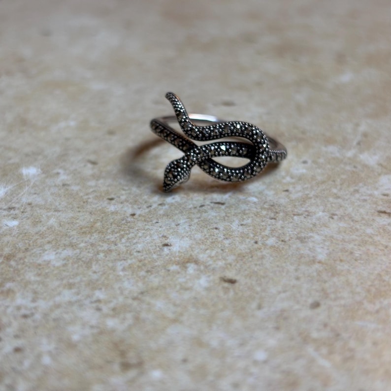 May include: A silver-toned snake ring with a detailed, textured design. The snake curves around the finger, with a head and tail that meet. The ring is set against a neutral background.