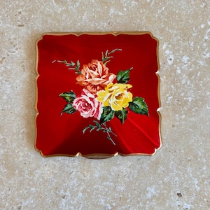 May include: A square compact mirror with a glossy red finish and scalloped edges. The centre features a detailed floral illustration of three roses in shades of orange, yellow, and pink, accompanied by green leaves and thin stems on a red background.