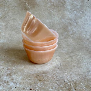 Puede incluir: Una pila de cuatro cuencos pequeños de vidrio color melocotón. Los cuencos tienen un borde festoneado y un acabado brillante. Los cuencos están apilados uno encima del otro, con el cuenco superior ligeramente inclinado.