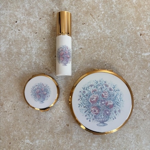 Peut inclure: Un atomiseur de parfum cylindrique avec un capuchon doré et un corps blanc orné d'un motif de bouquet floral, accompagné d'un petit poudrier rond et d'un plus grand, tous deux avec des bords dorés et des motifs floraux assortis sur fond blanc.