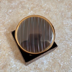 May include: A round compact mirror featuring a polished gold-tone metal frame and a smooth, dark brown stone inlay with vertical white banding. The compact is nestled within a square, dark-colored box with a textured, patterned exterior finish.