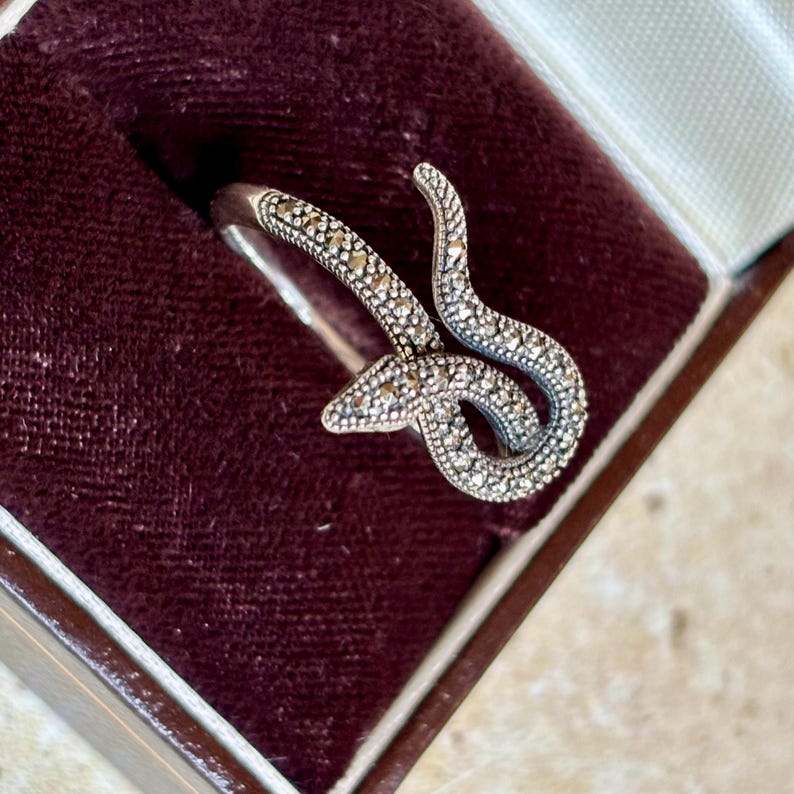 May include: A silver-toned ring in the shape of a snake, with a textured, gem-studded design. The ring is displayed in a burgundy velvet jewelry box. The snake's body curves and coils, creating a unique and eye-catching accessory.