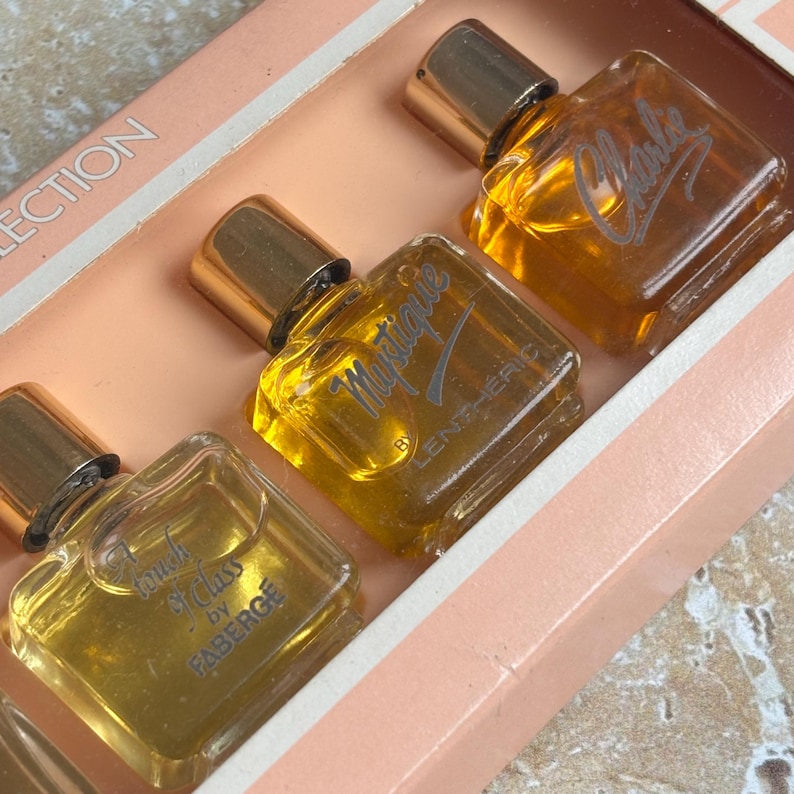 May include: A vintage perfume collection set in a pink box. The set includes three small, square glass bottles with gold-colored caps. The bottles are labeled "Charles", "Mystique", and "Touch of Class by Faberge". The perfume inside is amber-colored.
