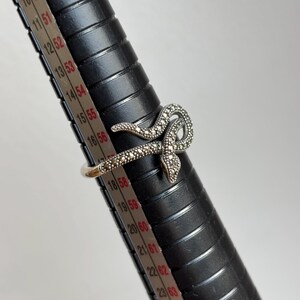 May include: A silver-toned snake ring with a detailed marcasite design. The snake wraps around the finger, with the head and tail overlapping. The ring is displayed on a black ring sizer with measurements in inches.