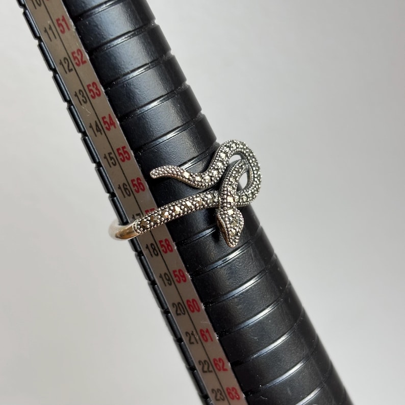 May include: A silver-toned snake ring with a detailed marcasite design. The snake wraps around the finger, with the head and tail overlapping. The ring is displayed on a black ring sizer with measurements in inches.