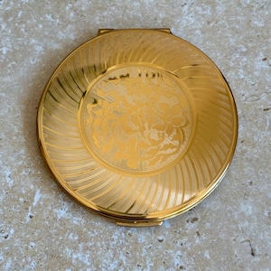May include: A round gold-tone metal compact mirror featuring a polished finish. The outer edge has a textured swirl pattern, while the center displays an engraved floral design. The compact is circular with a hinged closure at the top edge.