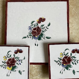 May include: A set of three square ceramic tiles with a floral design. The tiles have a white background with a burgundy border. The floral design features burgundy, yellow, blue, and green leaves.