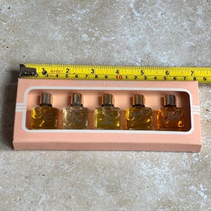 May include: A vintage fragrance collection set in a pink box. The set includes five small perfume bottles with gold caps, each containing a different amber-colored fragrance. The box is labeled "FRAGRANCE COLLECTION" and is measured with a tape measure.