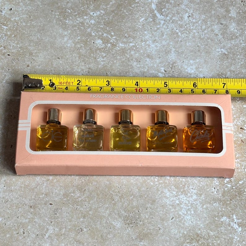 May include: A vintage fragrance collection set in a pink box. The set includes five small perfume bottles with gold caps, each containing a different amber-colored fragrance. The box is labeled "FRAGRANCE COLLECTION" and is measured with a tape measure.