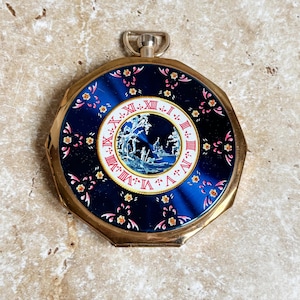 Pode incluir: Um relógio de bolso vintage com caixa dourada e mostrador azul escuro. O mostrador do relógio apresenta algarismos romanos e uma cena de paisagem detalhada. Padrões florais delicados em rosa e amarelo adornam o fundo azul.