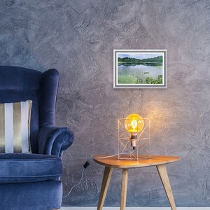 May include: A framed watercolor painting of a lake scene hangs on a textured gray wall. A blue armchair with a striped pillow sits to the left. A small wooden side table with a geometric lamp is to the right, creating a cozy interior design.