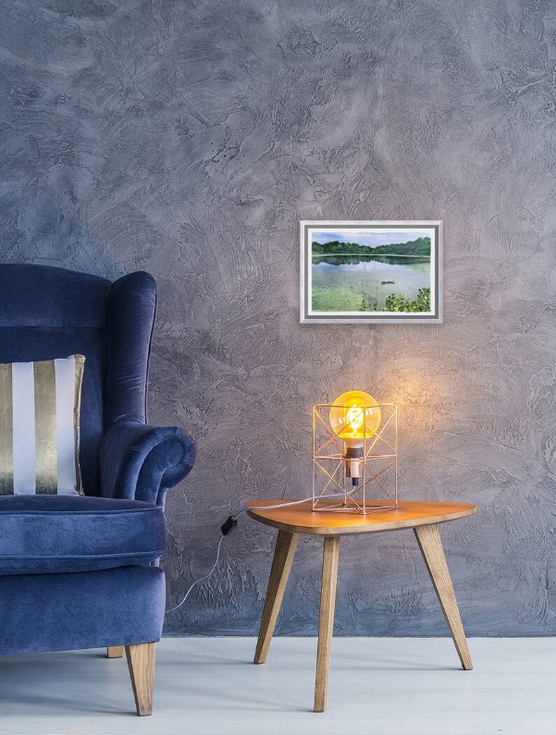 May include: A framed watercolor painting of a lake scene hangs on a textured gray wall. A blue armchair with a striped pillow sits to the left. A small wooden side table with a geometric lamp is to the right, creating a cozy interior design.