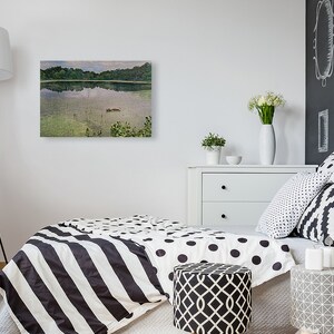 May include: A framed landscape painting depicts a serene lake scene with lush green trees reflected in the water. The artwork hangs on a white wall above a bed with black and white bedding. A white dresser and decorative pillows complete the room.