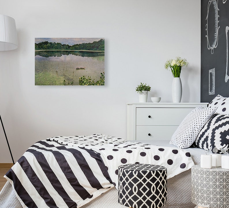 May include: A framed landscape painting depicts a serene lake scene with lush green trees reflected in the water. The artwork hangs on a white wall above a bed with black and white bedding. A white dresser and decorative pillows complete the room.