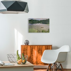May include: A landscape painting of a calm lake with trees on the far shore. The water reflects the sky and trees. The painting is displayed above a wooden cabinet. A modern white chair and a geometric pendant light are also in the room.