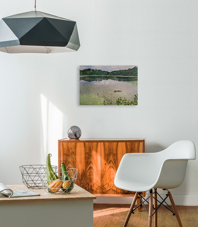 May include: A landscape painting of a calm lake with trees on the far shore. The water reflects the sky and trees. The painting is displayed above a wooden cabinet. A modern white chair and a geometric pendant light are also in the room.