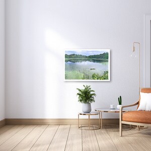 May include: A framed watercolor painting of a serene lake scene hangs on a white wall. The artwork depicts a calm lake with green vegetation and trees in the background. The room features a modern chair, side tables, and plants.