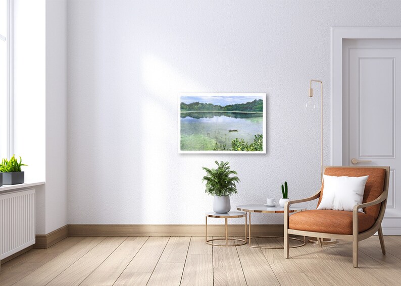 May include: A framed watercolor painting of a serene lake scene hangs on a white wall. The artwork depicts a calm lake with green vegetation and trees in the background. The room features a modern chair, side tables, and plants.
