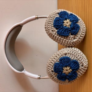 以下が含まれることがあります： 白いヘッドホンに、かぎ針編みで作った耳あてが付いています。耳あてはベージュで、青と黄色の花が飾られています。