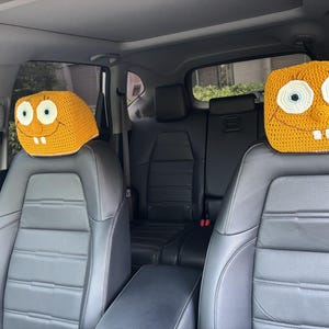 May include: Two yellow crocheted headrest covers shaped like a cartoon character's face, with white eyes, black pupils, and a smiling mouth with two teeth. The covers are on the front seats of a car.