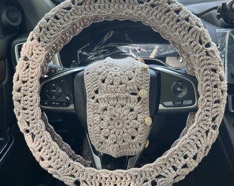 Op de afbeelding: Een beige gehaakte stuurwielhoes met een bijpassend gehaakt vierkant op het dashboard. De hoes heeft een delicaat, ingewikkeld patroon.