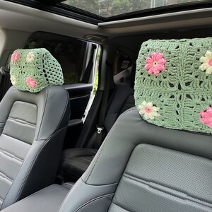 May include: Two green crocheted headrest covers with pink and white flowers. The covers are on the headrests of two black leather car seats.