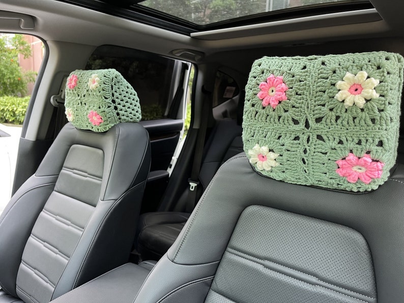May include: Two green crocheted headrest covers with pink and white flowers. The covers are on the headrests of two black leather car seats.