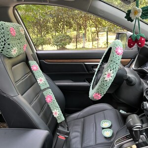 May include: Green crocheted car seat covers with pink flowers. The set includes a headrest cover, seatbelt cover, and steering wheel cover.